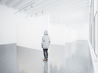 Obraz premium Photo of girl in empty modern gallery looking at the blank white canvas. Big windows, spotlights, concrete floor. Horizontal, mockup