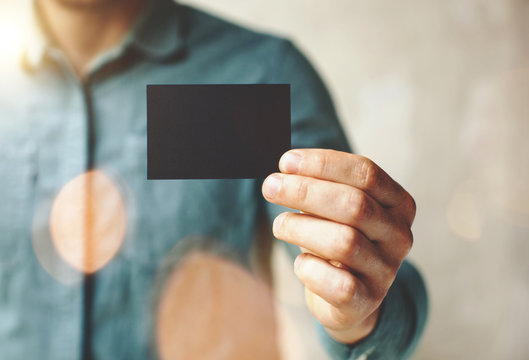 Man wearing blue jeans shirt and showing blank black business card. Blurred background. Horizontal mockup