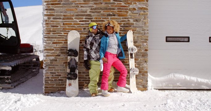 Cute couple in snowsuits standing next to upright snowboards while they lean against ski resort garage with tractor in thick snow on sunny day