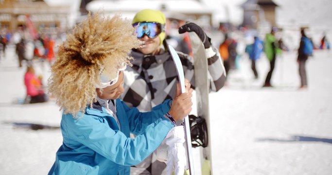Man in goggles flirting with smiling woman in blue jacket holding snowboard on crowded ski slope on sunny day