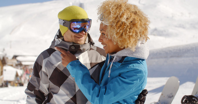 Adult young woman in blond curly hair looking at happy friend with goggles taking a break from snowboarding at ski resort surrounded by thick white snow outside.