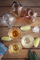 Three shots of Tequila with square bottles, lime and salt on a wooden surface viewed from above