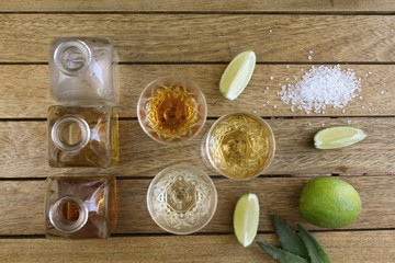 Three shots of alcoholic drinks next to square bottles with lime quarters and salt on a wooden surface viewed from above