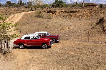 Kuba: Zwei rote US-amerikanische Oldtimer mit weißem Dach parken auf einem traditionellen Feld 