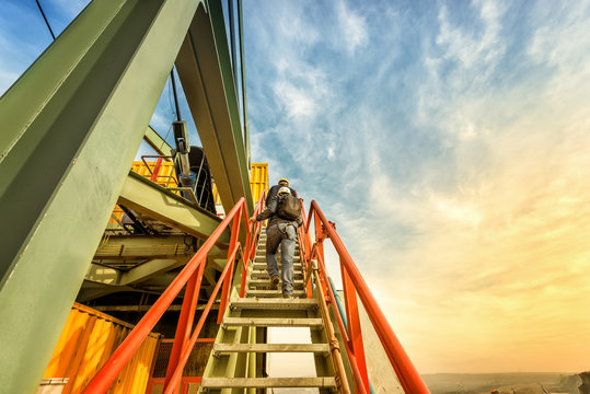 Two coal mine engineers are climbing on the steep stairs to the highest part of the huge coal digger machine. Beautiful and colorful sky in the background. Rear view. Look from below.