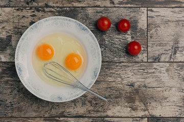 Two eggs with whisk and cherry tomatoes photographed from above on old grungy wooden table. Place for text.