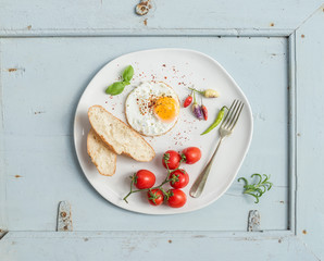 Breakfast set. Fried egg, bread slices, cherry tomatoes, hot peppers and herbs on white ceramic plate over light blue wooden backdrop, top view.