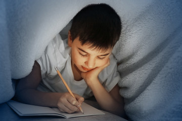 Little boy writing with pencil under the blanket