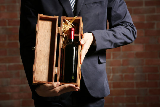 Man holding a bottle of red wine and glass in wooden box on brick wall background