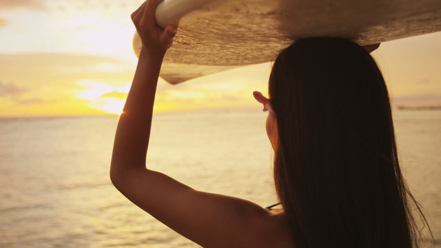 Young female surfer going surfing holding surfboard at sunset. Beautiful ethnic mixed race Asian Chinese / Caucasian woman sport model. Sun flare and colorful silhouette. Waikiki, Oahu, Hawaii, USA.