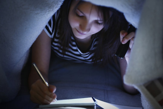 Beautiful girl writing in notebook with flashlight under the blanket