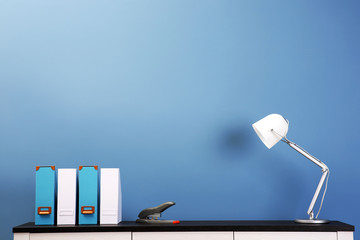 Wooden chest of drawers with folders, lamp and stapler on blue wall background