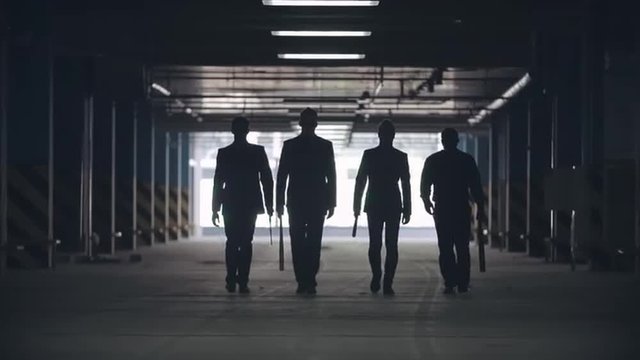 Black silhouettes of four men walking towards the camera in the car parking