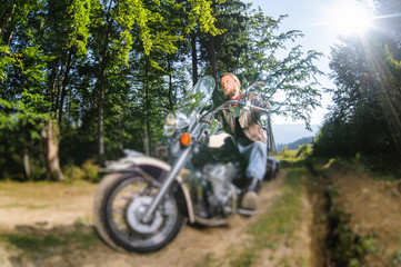 Fototapeta na wymiar Handsome biker with beard driving his cruiser motorcycle in the forest. Man is wearing leather jacket and blue jeans. Wide angle. Tilt shift lens blur effect
