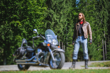 Handsome biker with long hair and beard standing near his custom made cruiser motorcycle. Biker is wearing leather jacket and sunglasses on sunny day. tilt shift soft effect