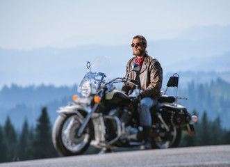 Man with beard in sunglasses, blue jeans and a leather jacket sitting on the travel motorbike and relaxing. Sunny day in the mountains. Tilt shift soft effect