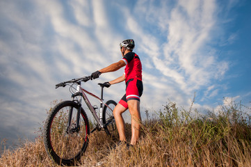 Fototapeta na wymiar Mountain Bike cyclist in red sportswear and helmet resting with his bike against blue sky. Biker is looking into distance. Low angle view.