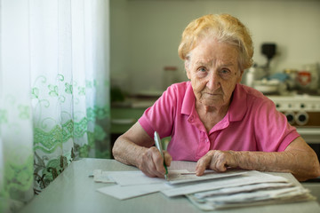 The old woman fills out utility bills sitting in the kitchen.