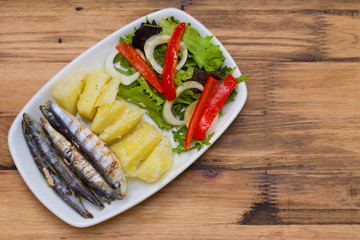 fried sardines with boiled potato and salad on white plate on brown wooden background