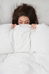 Portrait of a young woman lying in bed hiding under duvet.