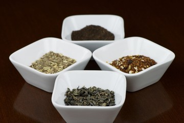Four sorts of dry tea leaves in square plates on brown wooden table with reflection