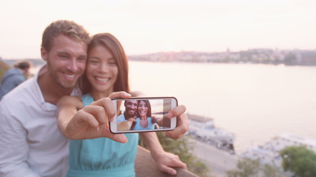 Couple taking selfie self portrait photo picture in Stockholm. Candid Scandinavian man and Asian woman looking at old town cityscape sunset view from Monteliusvagen overlooking Gamla Stan the old city