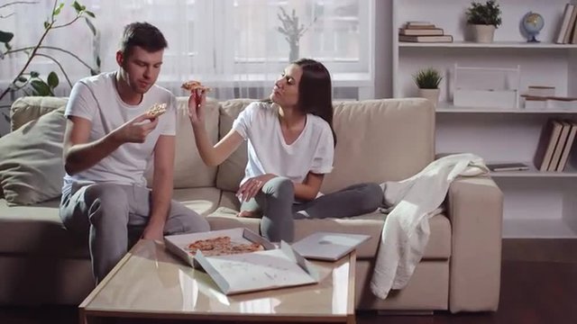 Young man and woman sitting on the couch together and eating pizza: girlfriend feeding her boyfriend with slice of pizza and smiling