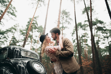 Stylish Loving wedding couple kissing and hugging in a pine forest near retro car