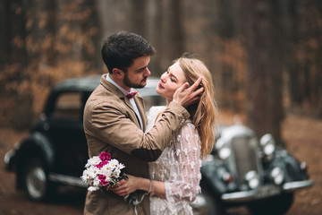 Stylish Loving wedding couple kissing and hugging in a pine forest near retro car
