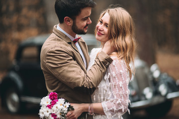 Stylish Loving wedding couple kissing and hugging in a pine forest near retro car