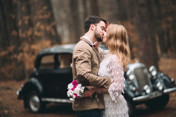 Stylish Loving wedding couple kissing and hugging in a pine forest near retro car