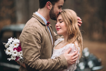 Stylish Loving wedding couple kissing and hugging in a pine forest near retro car