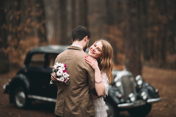 Stylish Loving wedding couple kissing and hugging in a pine forest near retro car