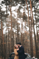 Stylish Loving wedding couple kissing and hugging in a pine forest near retro car