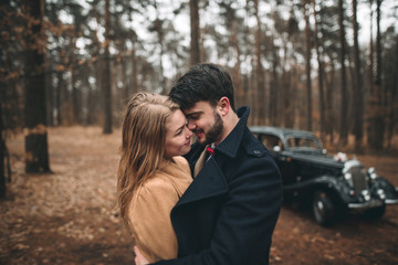 Stylish Loving wedding couple kissing and hugging in a pine forest near retro car
