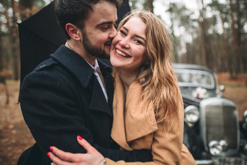 Stylish Loving wedding couple kissing and hugging in a pine forest near retro car