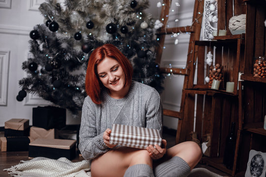 fashion interior photo of beautiful young woman with red hair and charming smile, wears cozy knitted cardigan, posing beside Christmas tree and presents with chocolate cake with asterisks