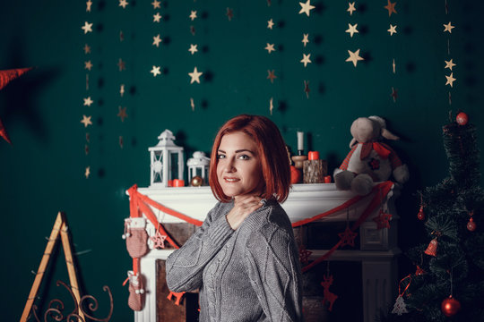 fashion interior photo of beautiful young woman with red hair and charming smile, wears cozy knitted cardigan, posing beside Christmas tree and presents with chocolate cake with asterisks