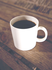 A plain white cup of black coffee on a wooden and antique box. Image has a vintage effect applied.