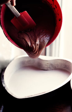 Pouring chocolate cake batter from a red mixing bowl into a prepared heart shaped baking pan
