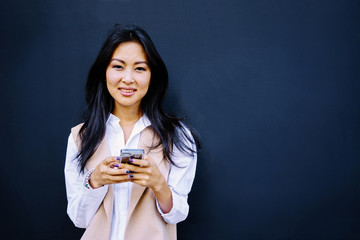 beautiful young Asian businesswoman typing on the smart-phone on the background of black wall with copy space for your text or design.
