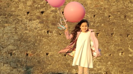 girl in a white dress and a pink coat with bright multi-colored balloons in the sun against the backdrop of the medieval walls