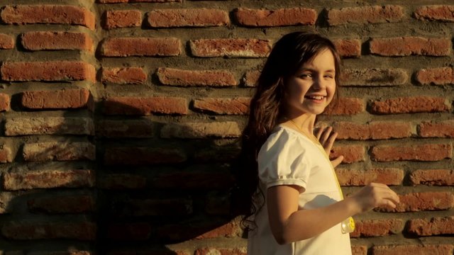 beautiful little girl near brick wall looking at the camera and playing with her hair