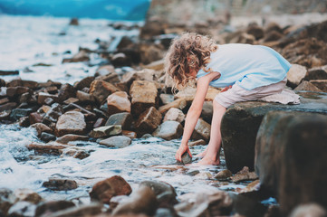happy kid girl playing with water on the stone beach. Cozy vacation on sea, traveling with kids.