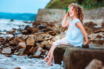 happy kid girl playing with water on the stone beach. Cozy vacation on sea, traveling with kids.