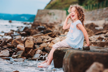 happy kid girl playing with water on the stone beach. Cozy vacation on sea, traveling with kids.