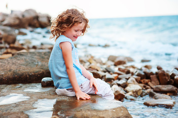happy kid girl playing with water on the stone beach. Cozy vacation on sea, traveling with kids.