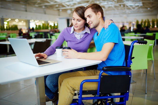 Amorous girl embracing her boyfriend in wheelchair while networking in cafe