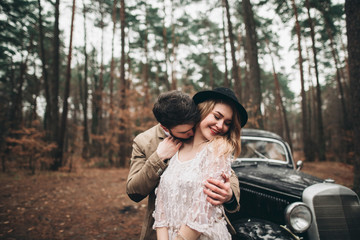 Gorgeous newlywed bride and groom posing in pine forest near retro car in their wedding day