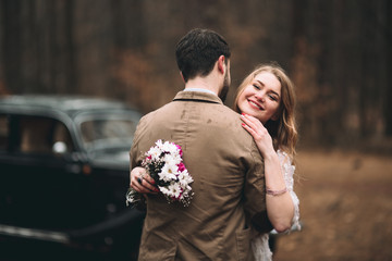Gorgeous newlywed bride and groom posing in pine forest near retro car in their wedding day